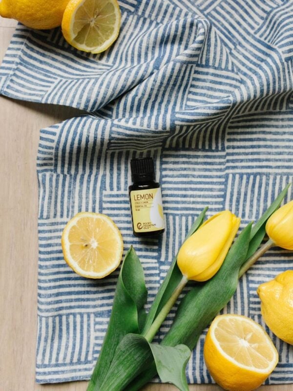 A bottle of lemon essential oil from Rocky Mountain Oils laying on a blue cloth. Sliced lemons and yellow flowers next to the bottle.