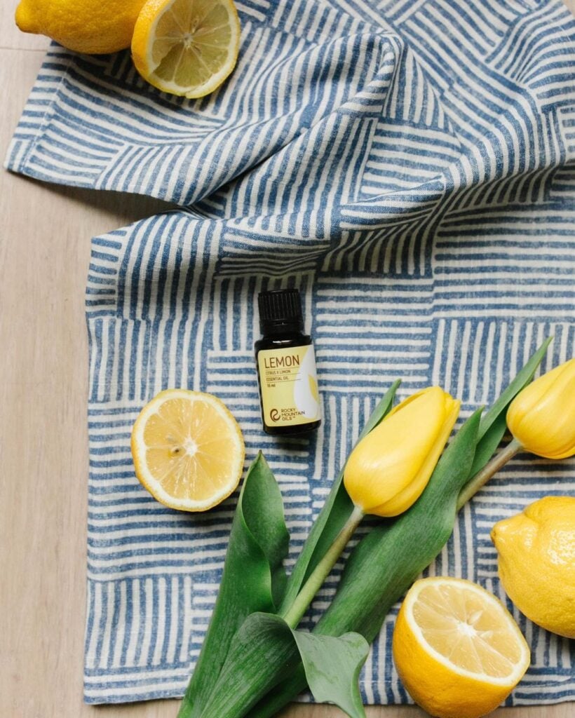A bottle of lemon essential oil from Rocky Mountain Oils laying on a blue cloth. Sliced lemons and yellow flowers next to the bottle.