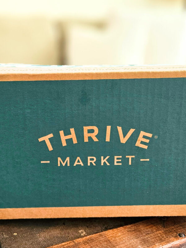 A cardboard box with Thrive Market printed on a green background. The box is sitting on top of a trunk with a couch, pillows, and plants in the background.