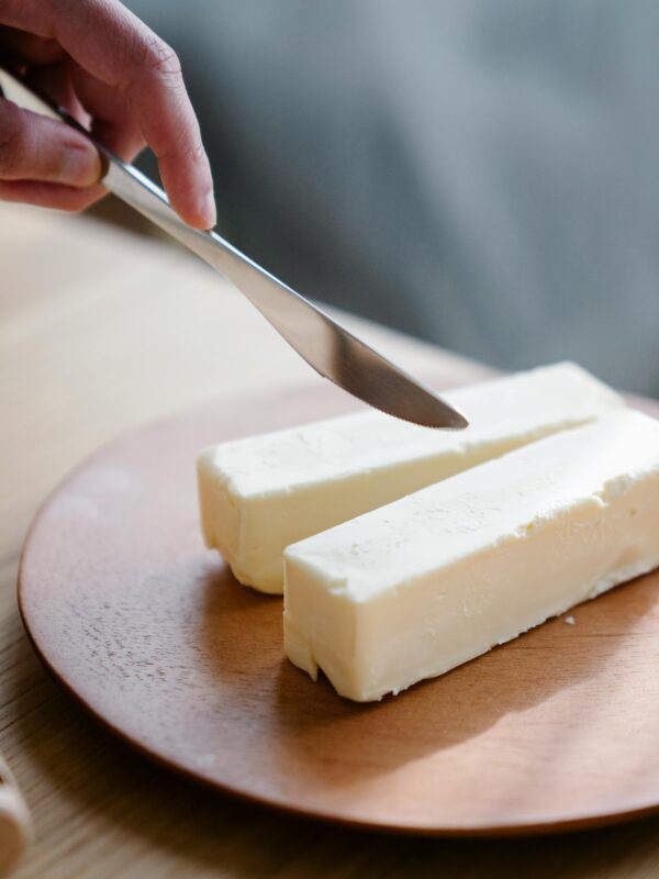 A hand holding a knife, ready to slice butter sticks on a wooden plate indoors. Butter, animal fats.