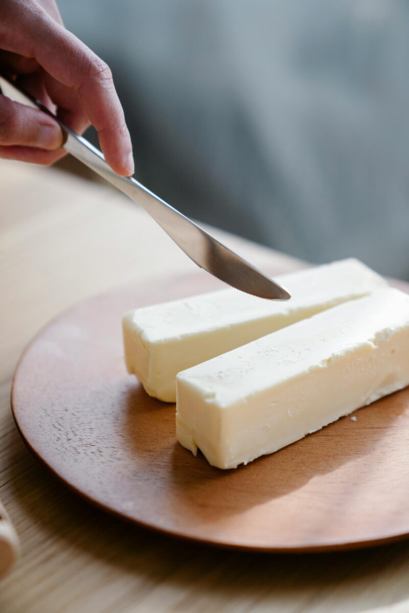 A hand holding a knife, ready to slice butter sticks on a wooden plate indoors. Butter, animal fats.