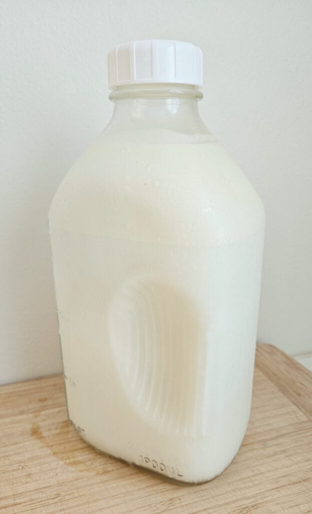 Half gallon milk jar full of raw milk. White lid. Sitting on top of a wooden cutting board.