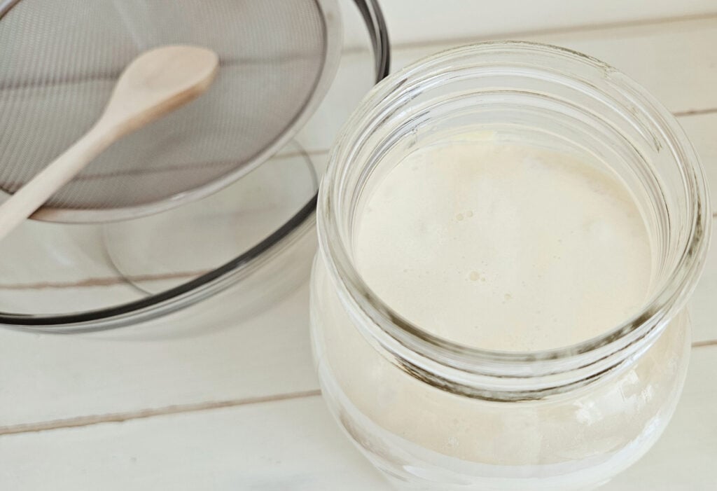 Top view of a jar of milk kefir sitting next to a glass bowl with a fine mesh strainer and wooden spoon on top.