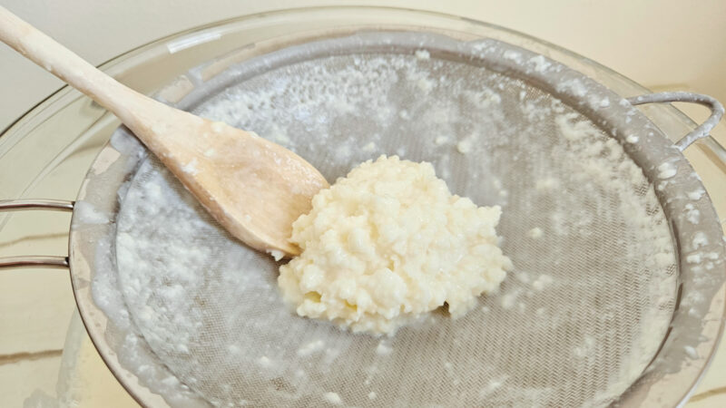 White/yellow milk kefir grains sitting on top of a stainless steel fine mesh strainer with a wooden spoon next to them. Strainer is resting on top of a glass bowl.