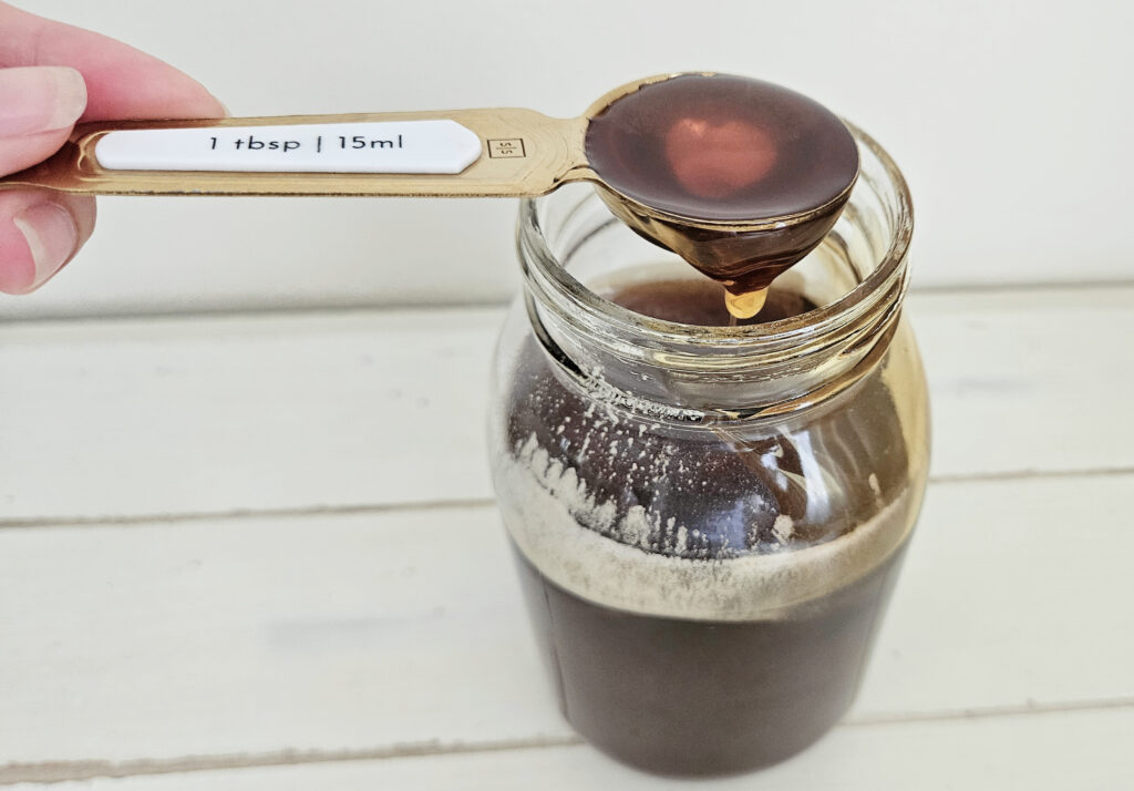 Woman hold a gold and white measuring spoon. One tablespoon of local raw honey dripping down from the measuring spoon into the mason jar full of dark brown honey.