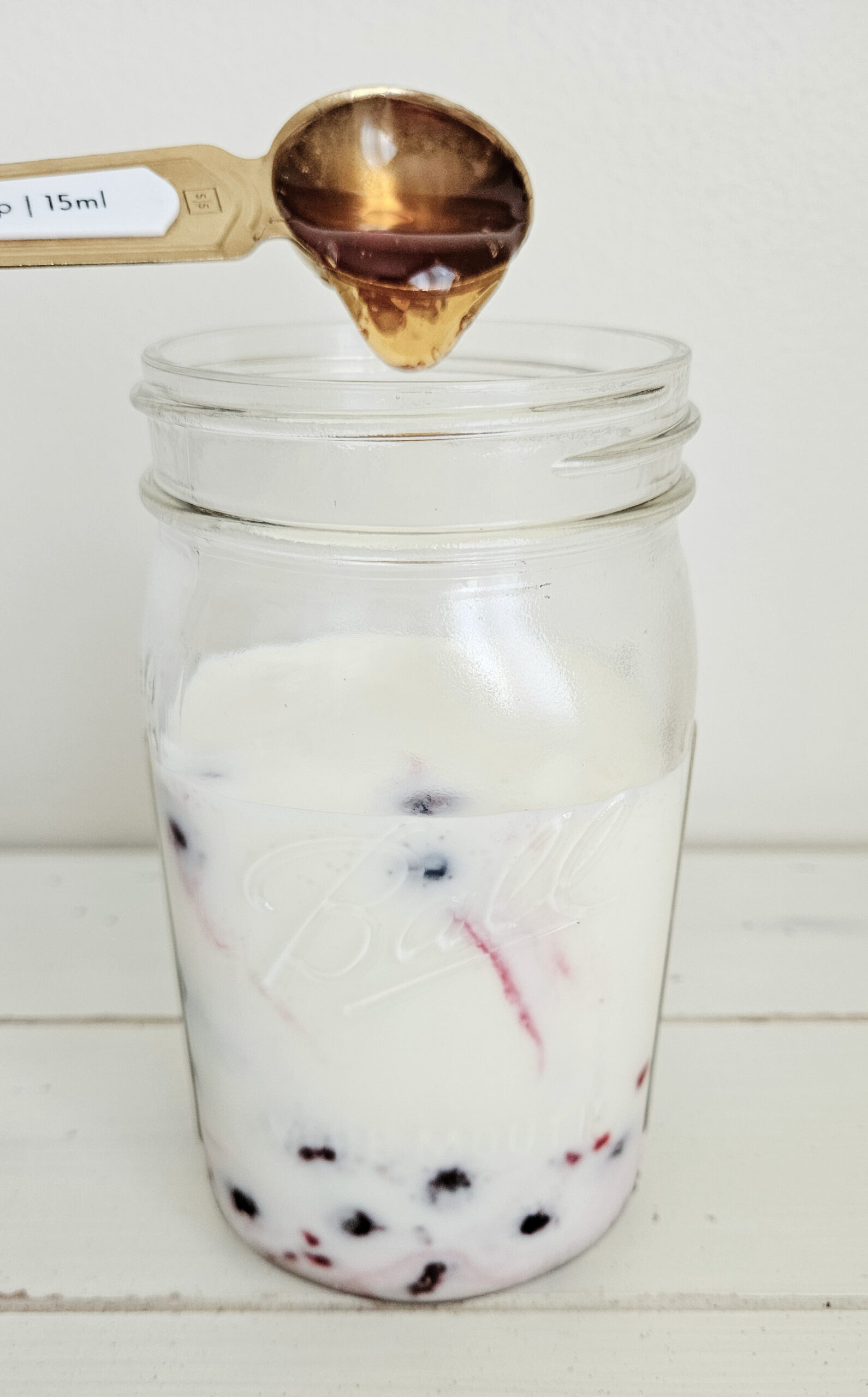 One tablespoon gold and white measuring spoon pouring raw local honey into a quart size mason jar filled with milk kefir and frozen mixed berries.