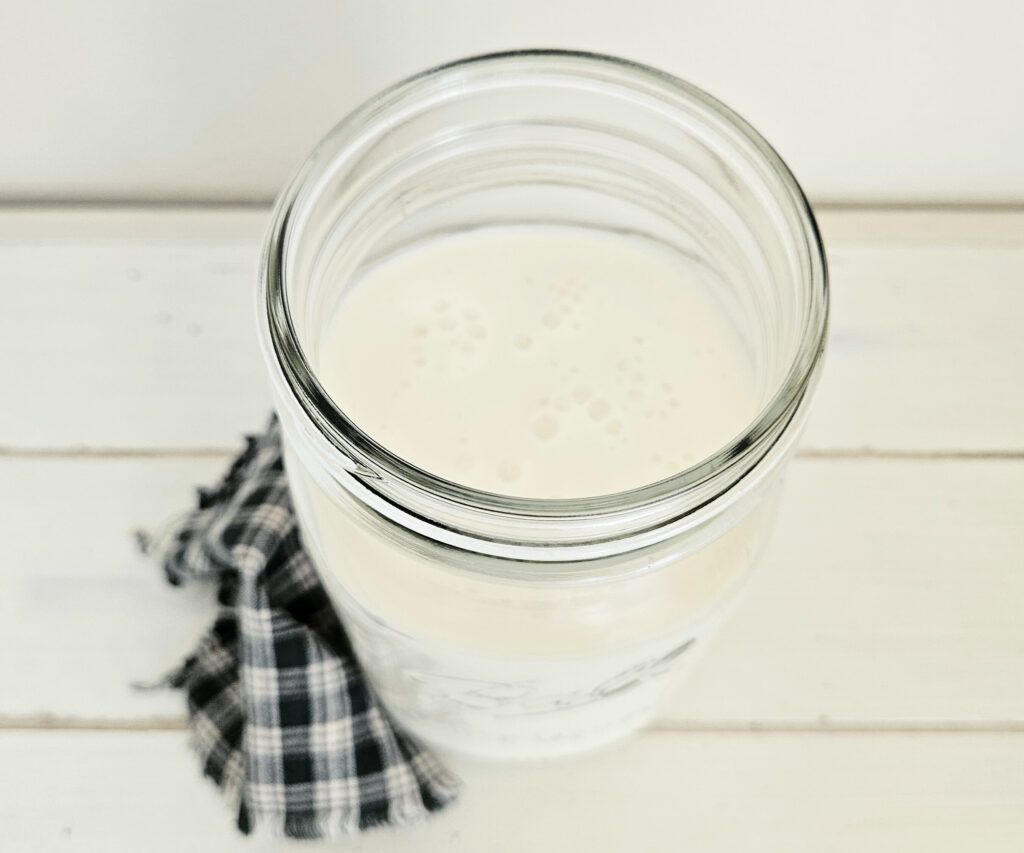 Aerial view of a wide mouth mason jar full of white bubbly kefir. No lid on top. Green checkered cloth on table next to jar.