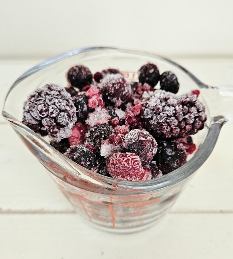 Glass measuring cup filled to the top with frozen mixed berries. Berries are purple and red. Frost is present on the berries.