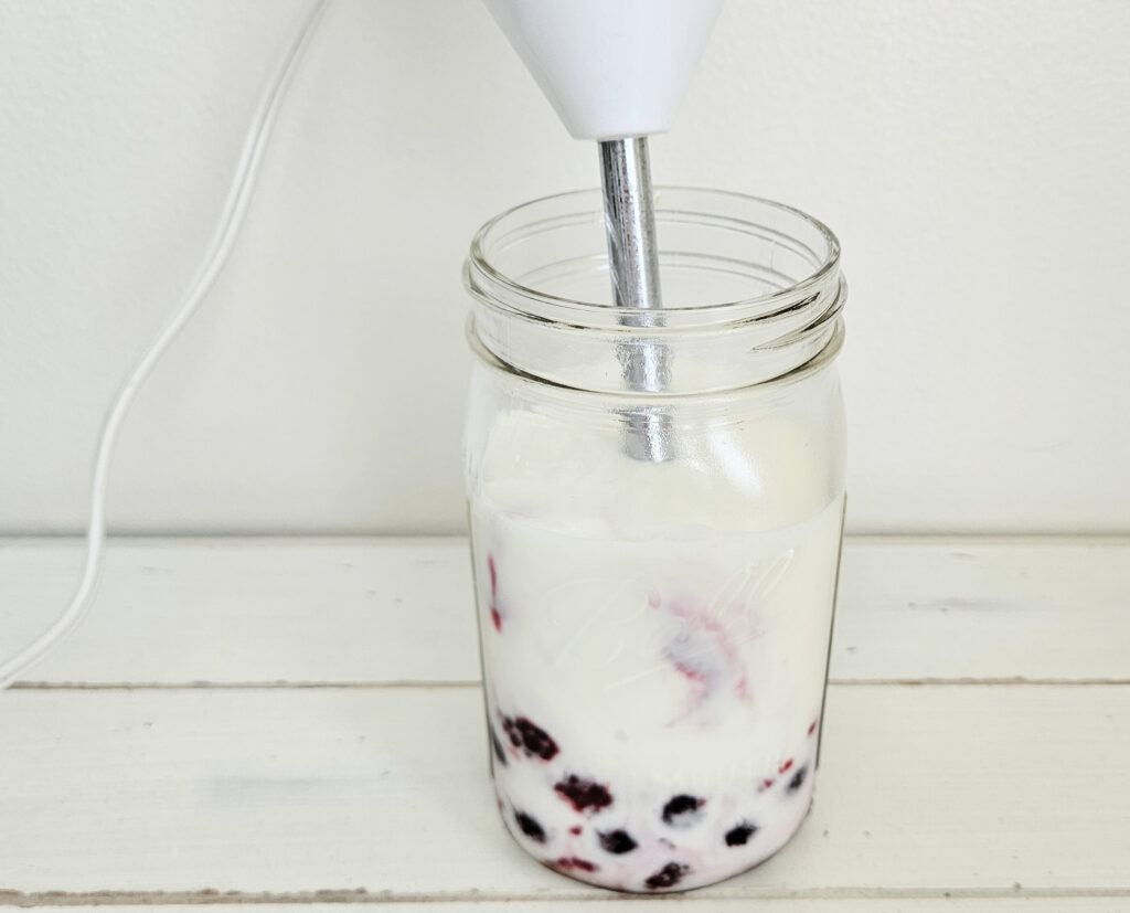 White and stainless steel immersion blender inside of a quart size mason jar. Inside the mason jar is milk kefir and frozen mixed berries.