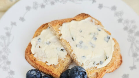 Kefir cheese with chives mixed in spread on two pieces of toast. Sitting on top of a white plate with a gray floral border. Blueberries in pile next to toast. Bowl of kefir cheese and a knife behind the plate.