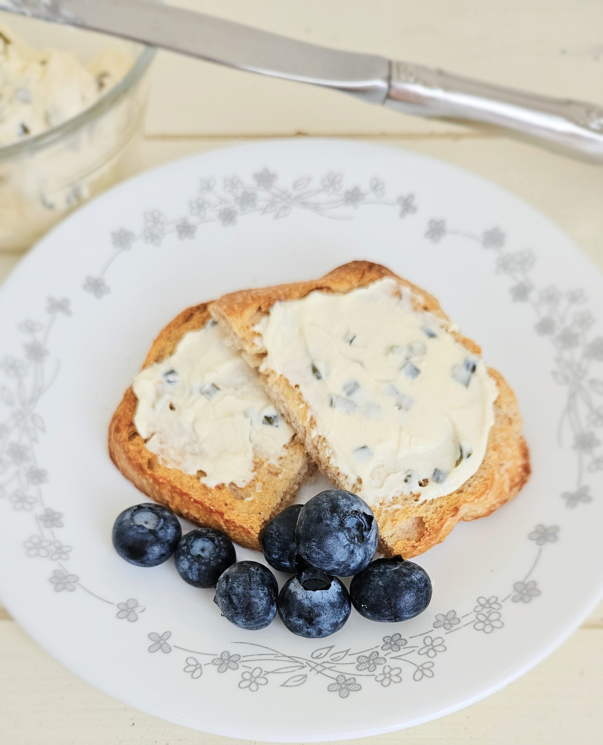 Kefir cheese with chives mixed in spread on two pieces of toast. Sitting on top of a white plate with a gray floral border. Blueberries in pile next to toast. Bowl of kefir cheese and a knife behind the plate.
