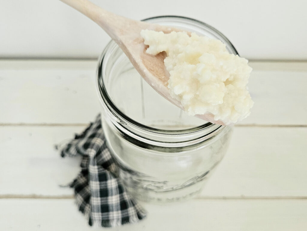 Pale yellow milk kefir grains sitting on top of a light colored wooden spoon. The spoon is resting on top of a wide mouth mason jar. There is a green and white checkered cloth sitting on the table next to the mason jar.