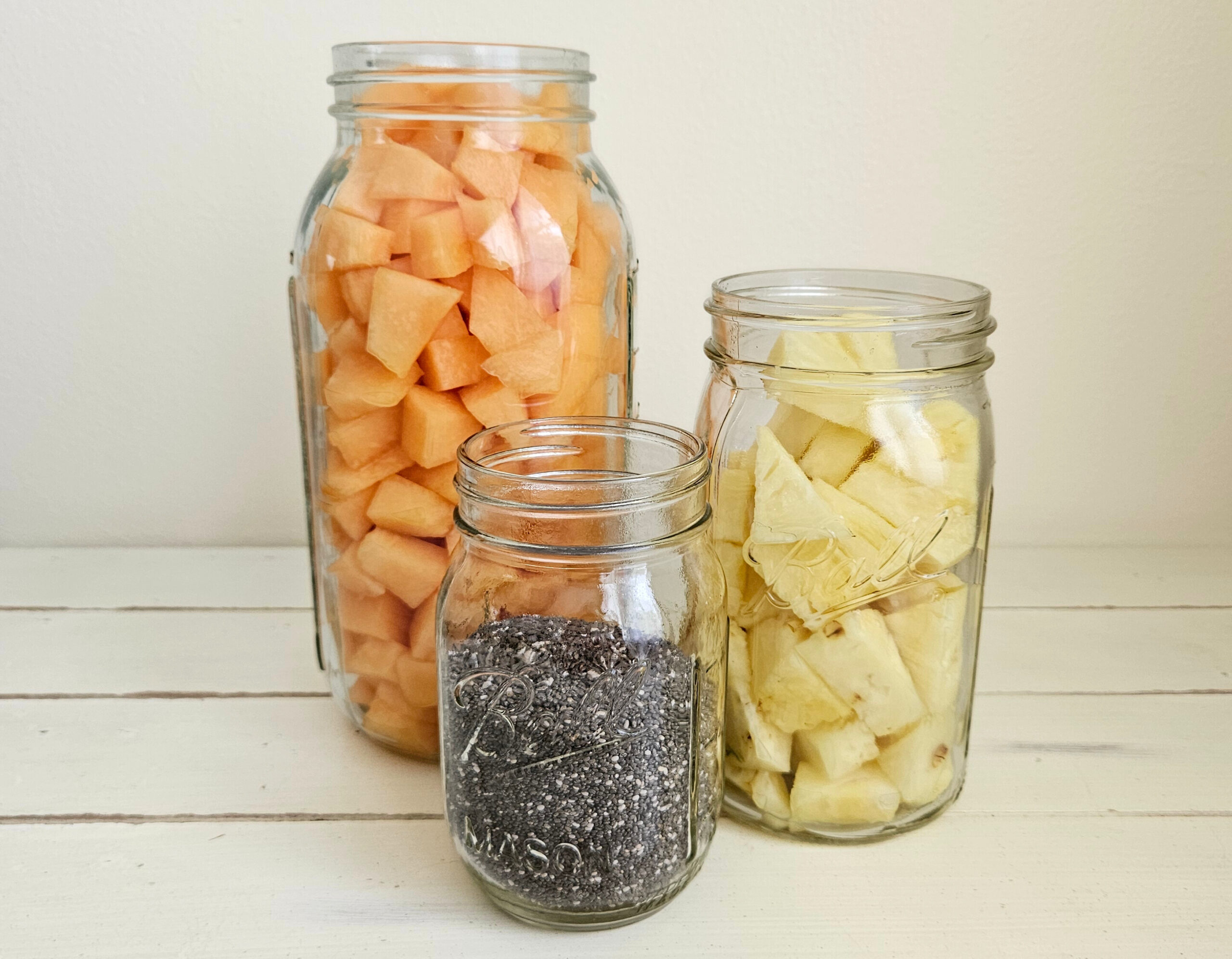 Half gallon mason jar filled with cut up cantaloupe. Quart size mason jar filled with cut up pineapple. Pint size mason jar filled with chia seeds. All toppings used for kefir smoothie bowls.