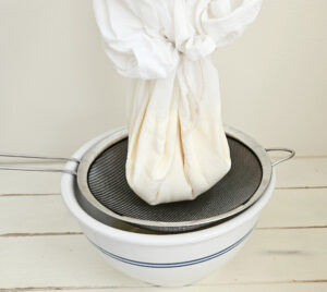 A white flour sack towel tied together with twine containing milk kefir curds. Whey dripping down onto fine mesh strainer and into an off white ceramic bowl.