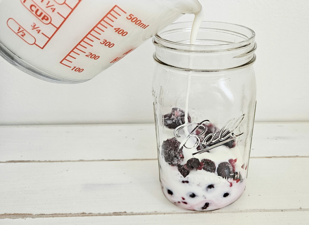 A wide mouth quart size mason jar with frozen mixed berries at the bottom. A glass measuring cup with red lettering on the side pouring 2 cups of milk kefir into the mason jar. The milk kefir and berries are mixed together in the jar.