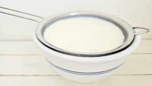 Milk kefir straining through a fine mesh strainer into an off white ceramic bowl with two blue lines as the outside bowl design.