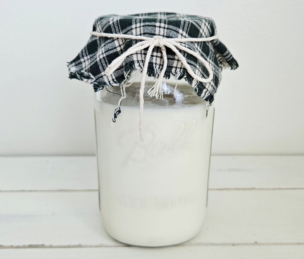 Wide mouth mason jar filled with white milk kefir. The top of the mason jar is covered with a green and white checkered cloth and secured with twine tied in a bowtie.