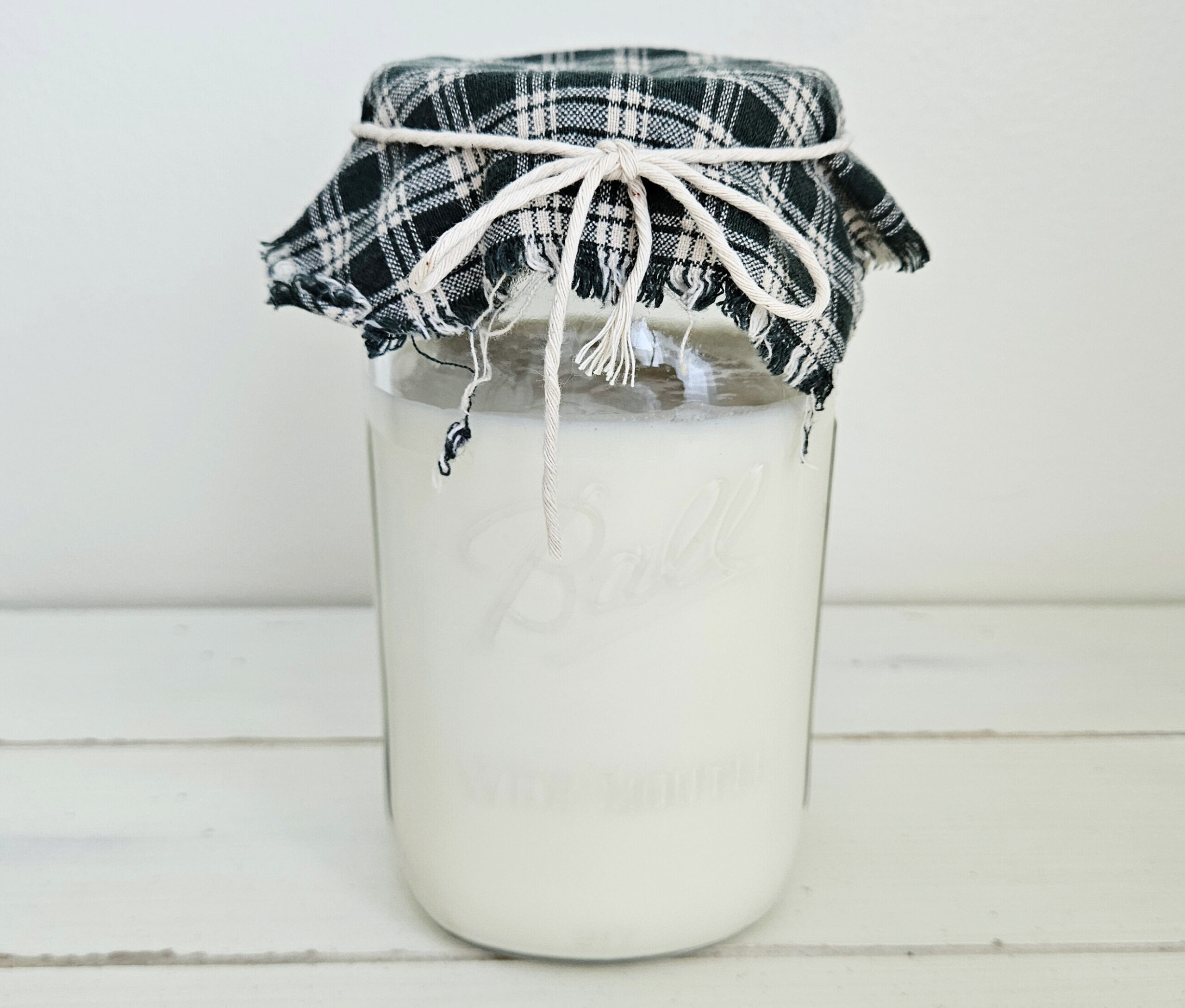 Wide mouth mason jar filled with white milk kefir. The top of the mason jar is covered with a green and white checkered cloth and secured with twine tied in a bowtie.