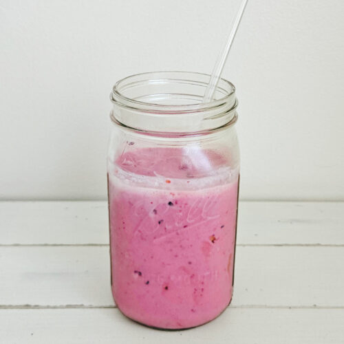 Mixed berry kefir smoothie in a glass mason jar with a glass straw sticking out. The smoothie is a bright magenta color. The mason jar is sitting on a cream colored table with a white wall background.