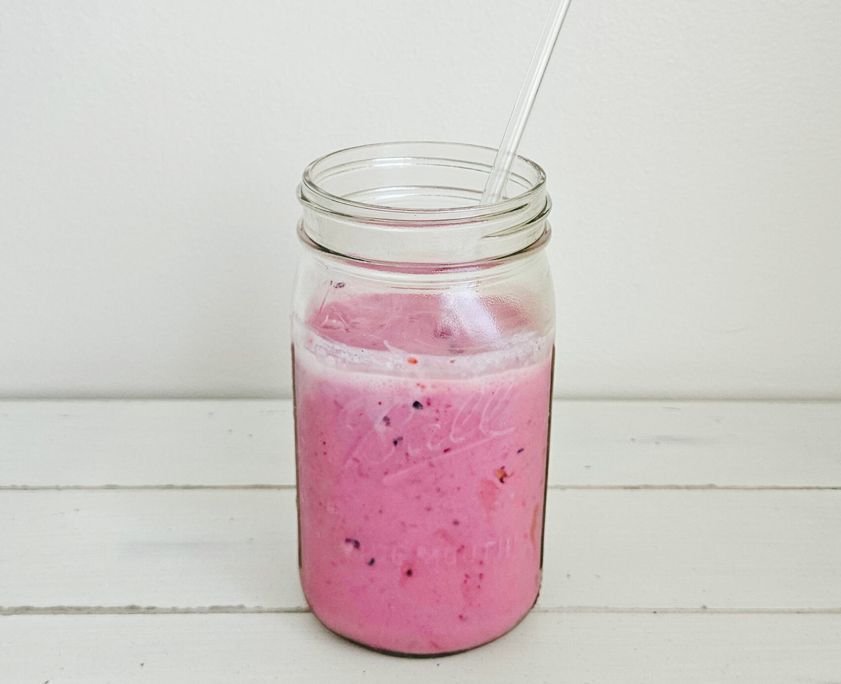 Mixed berry kefir smoothie in a glass mason jar with a glass straw sticking out. The smoothie is a bright magenta color. The mason jar is sitting on a cream colored table with a white wall background.