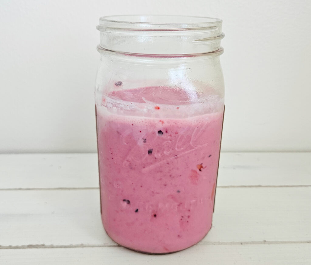 A wide mouth quart size mason jar filled with a mixed berry kefir smoothie. The smoothie is a bright magenta with red and purple spots throughout.