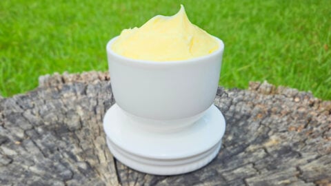 Bright yellow homemade butter in a white butter crock. Butter is higher than the rim of the crock. The crock is sitting on a tree stump. Green grass in the background.