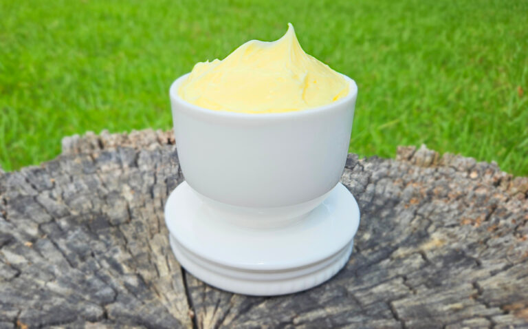 Bright yellow homemade butter in a white butter crock. Butter is higher than the rim of the crock. The crock is sitting on a tree stump. Green grass in the background.