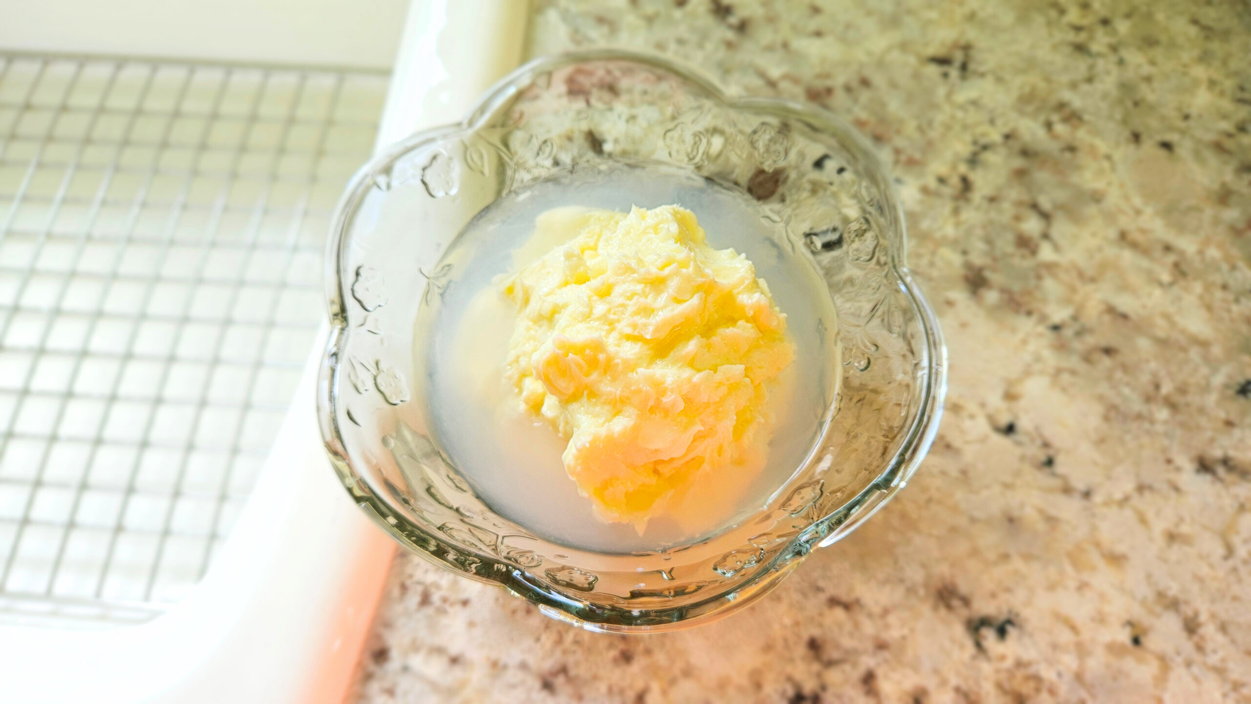 Homemade butter in a glass bowl sitting on top of a kitchen counter. Granite countertop and white sink. Water inside the bowl with the butter.