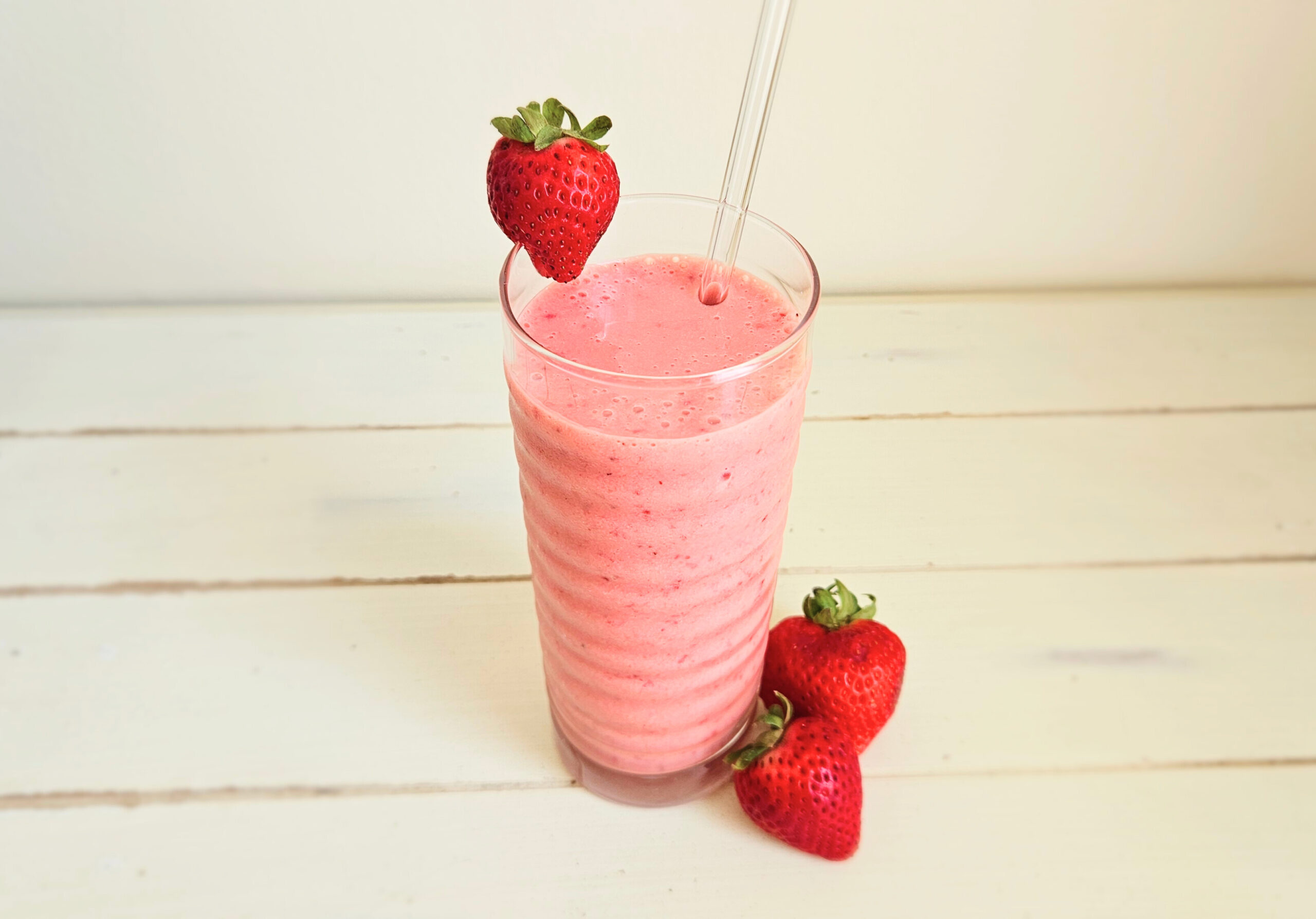 Strawberry banana kefir smoothie in a tall glass with a glass straw sticking out. Sliced strawberry on rim of glass. Two strawberries sitting next to glass. Mixed in is ancient nutrition multi collagen protein powder.