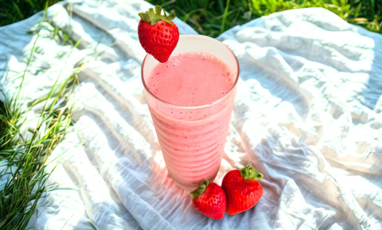 Pink colored strawberry banana kefir smoothie in a tall clear glass. Sitting on top of a white blanket. Blanket is on the grass. One strawberry on the rim of the glass. Two strawberries on the blanket next to the glass.