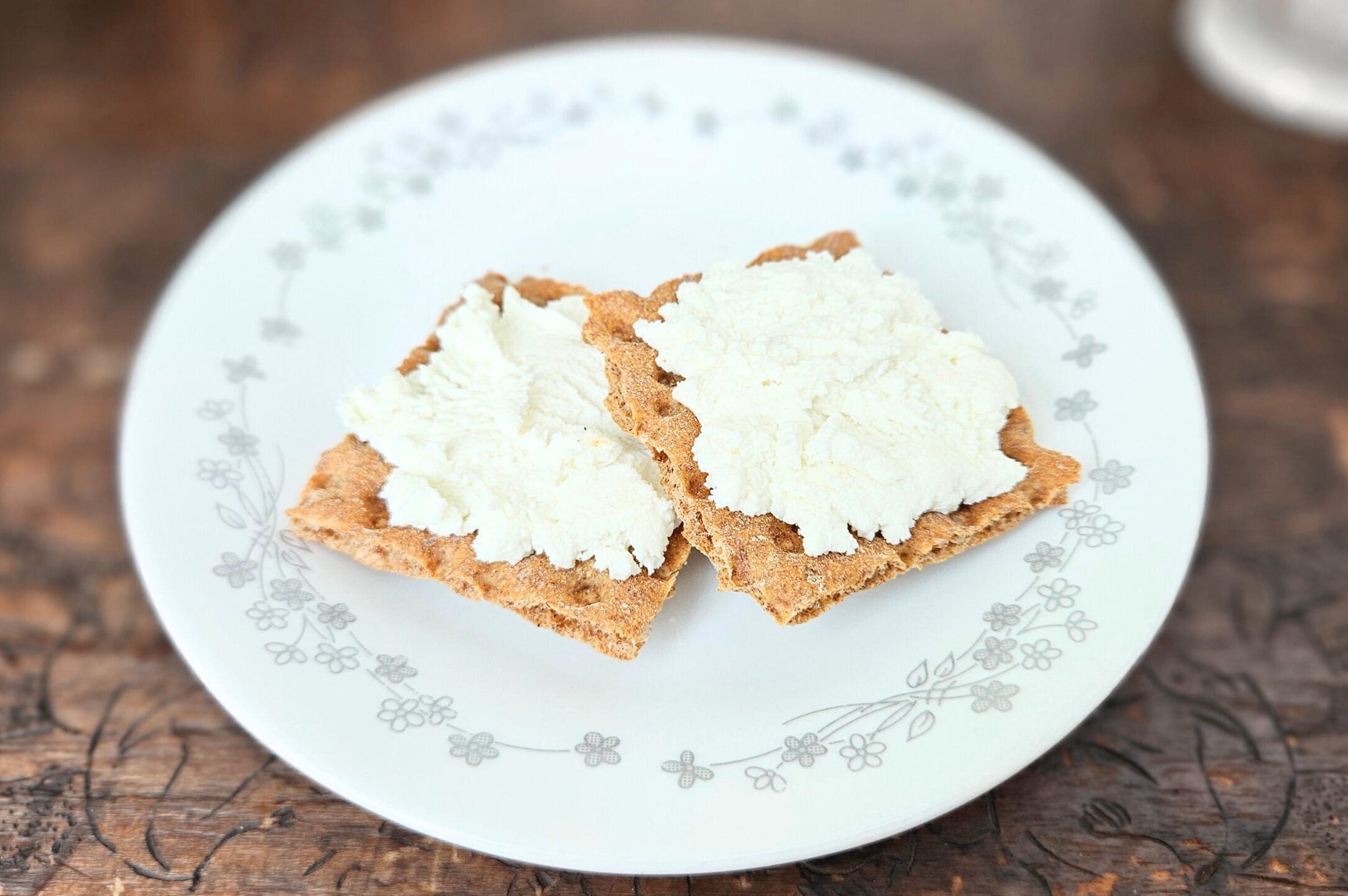 Two crackers with cream cheese on a white plate with a gray floral border. Sitting on top of a brown wooden trunk with plants in the background.