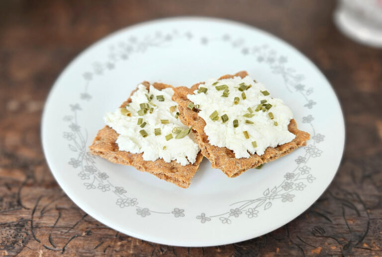 A white plate with a gray floral border with two crackers topped with cream cheese and chives. Sitting on top of a brown trunk.