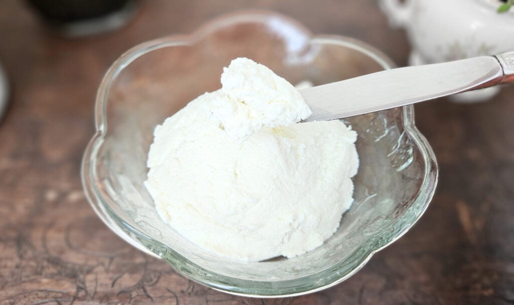Glass bowl of cream cheese with cream cheese on a silver knife hovering above. Sitting on a brown trunk.