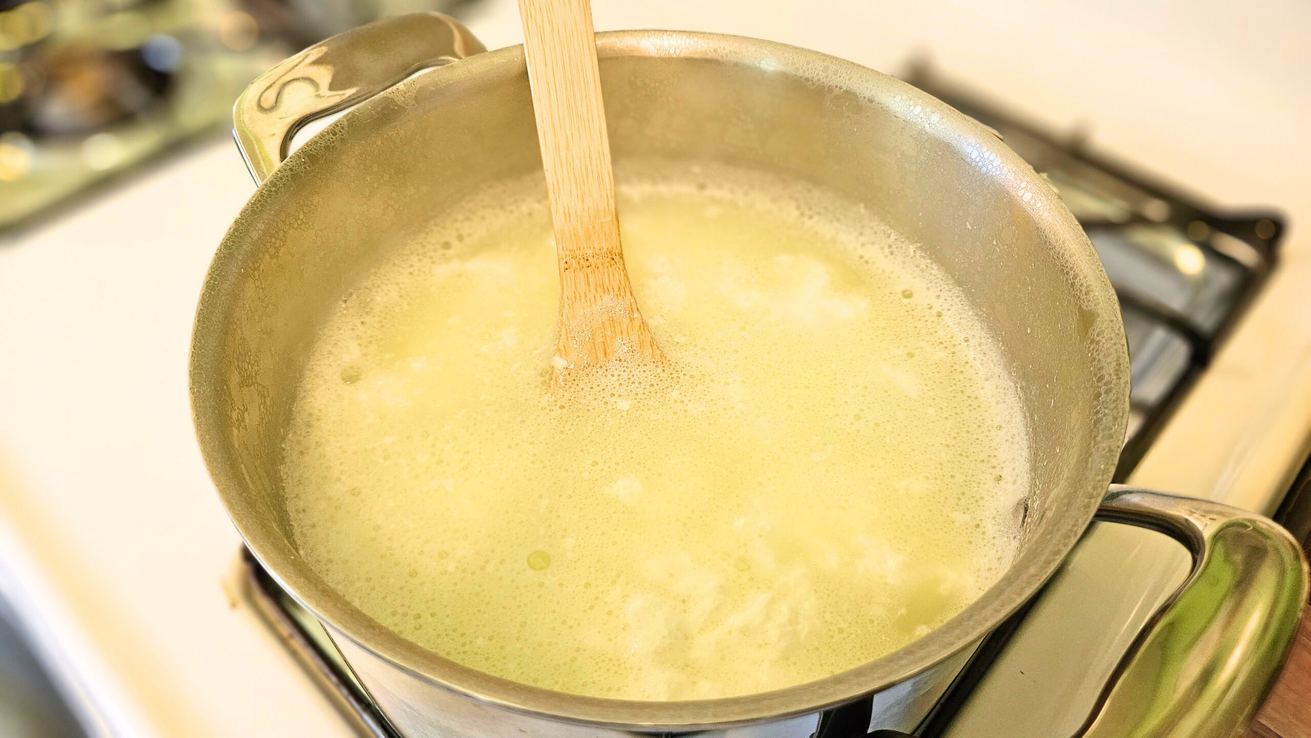 Stainless steel pot filled with curds and whey on a white stovetop. Wooden spoon in the pot.