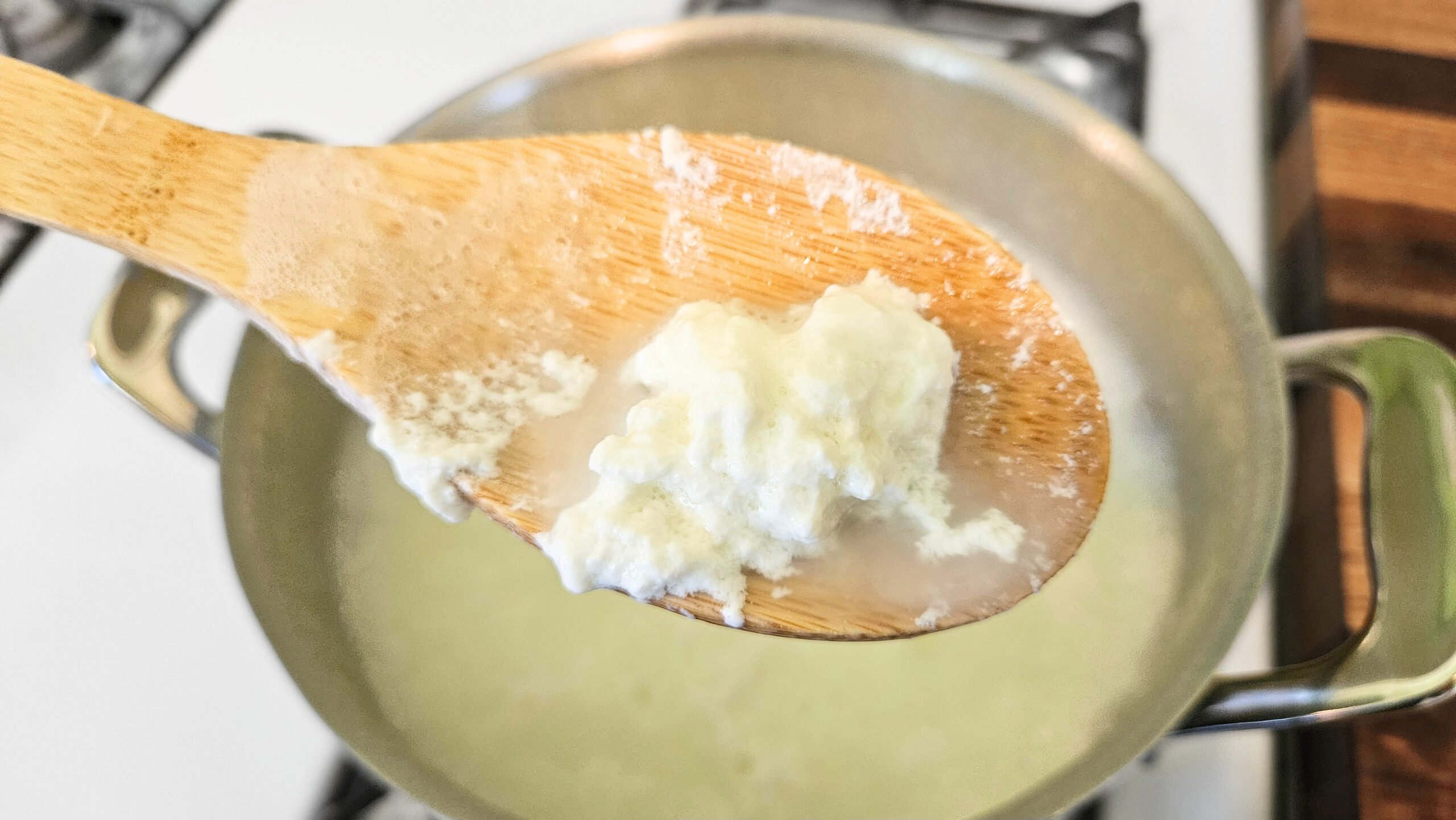 White curds on top of a wooden spoon with a pot full of curds and whey behind it.