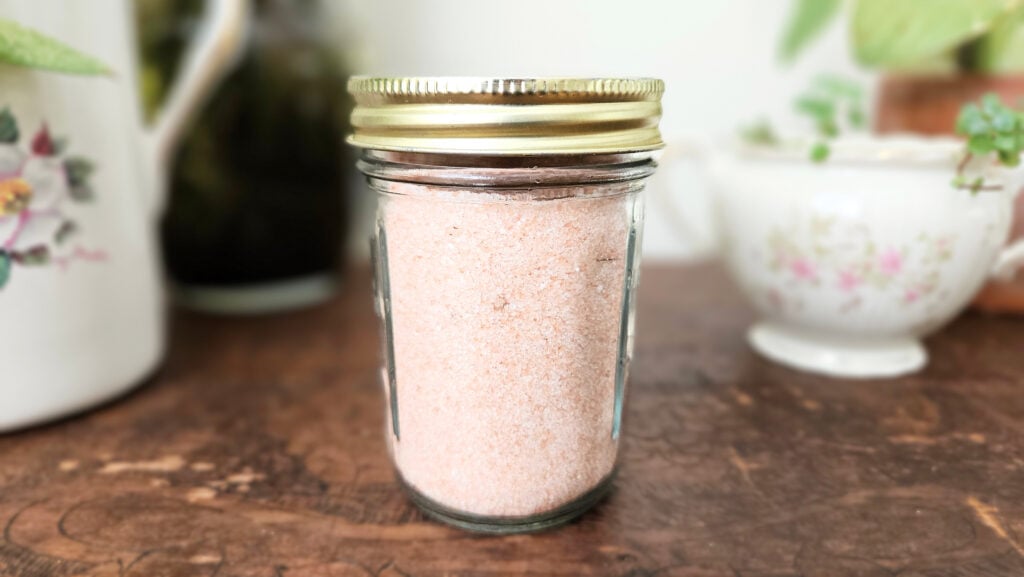 A mason jar filled with himalayan pink salt. Sitting on top of a brown wooden trunk with plants in the background.
