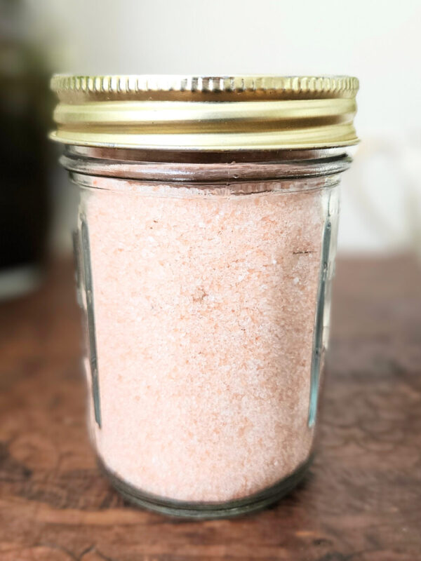 A mason jar filled with himalayan pink salt. Sitting on top of a brown wooden trunk with plants in the background.