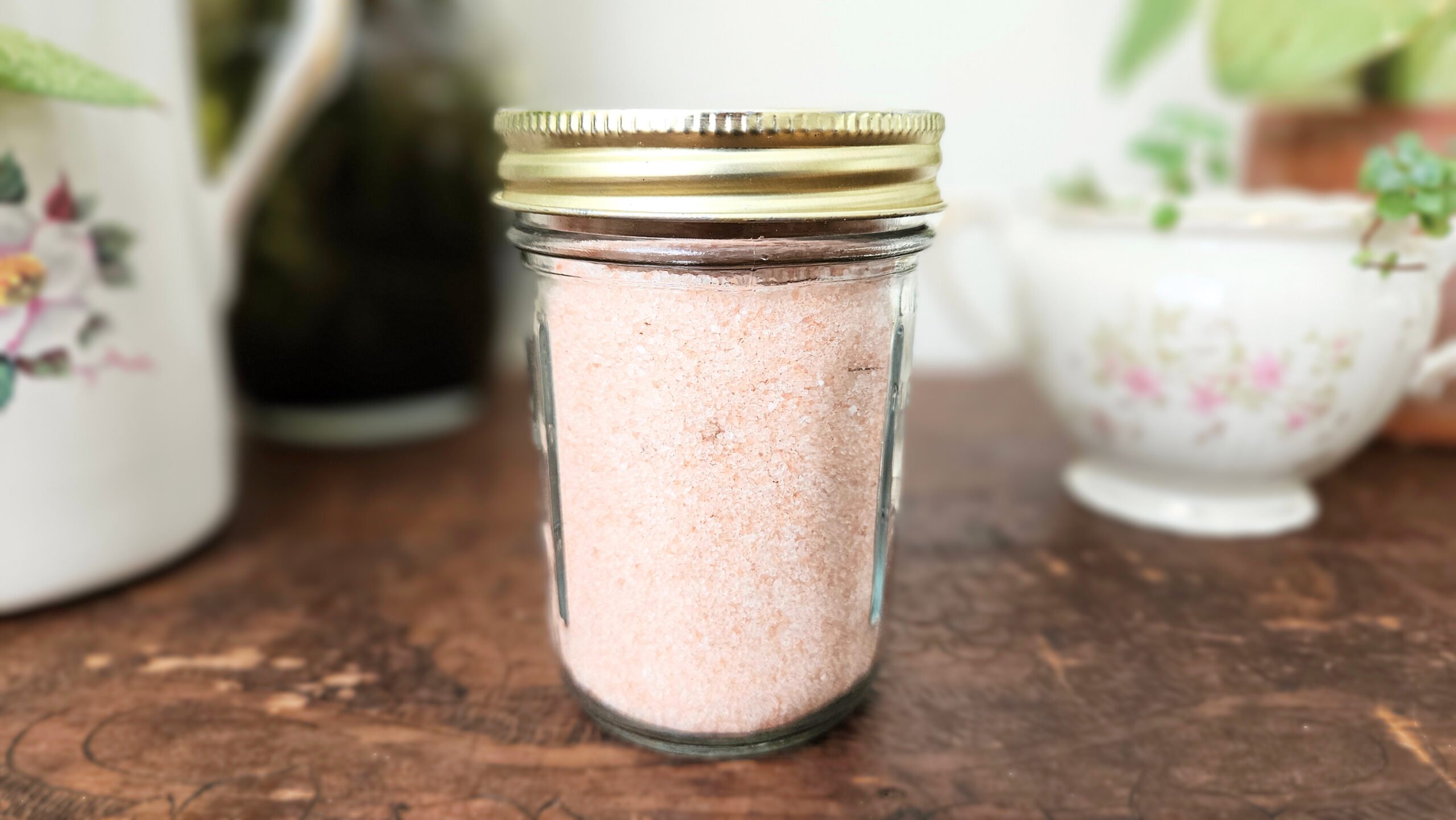 A mason jar filled with pink himalayan salt. Sitting on top of a brown wooden trunk with plants in the background.