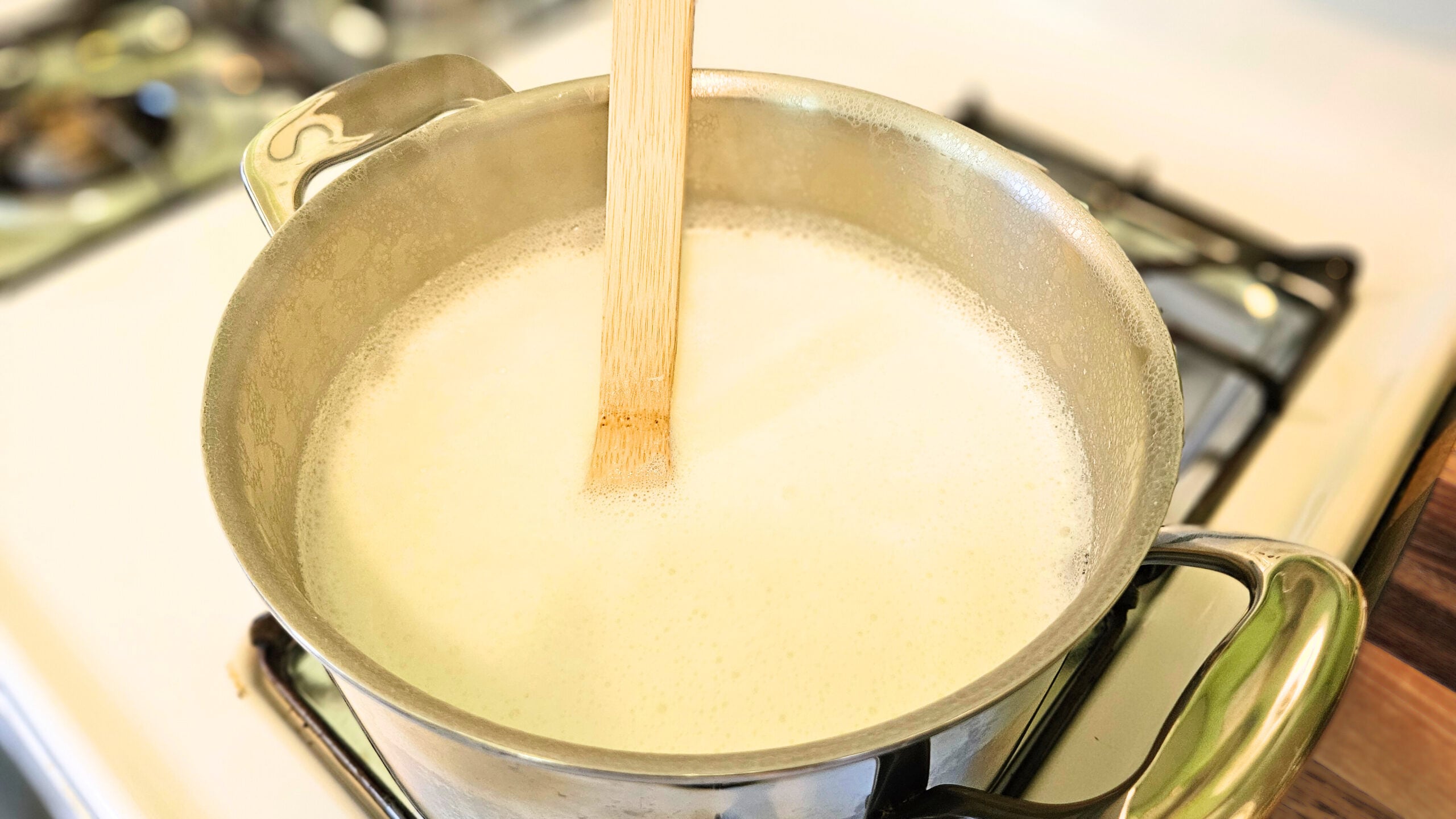 A stainless steel pot on top of a white stove filled with raw milk. Wooden spoon in the pot.
