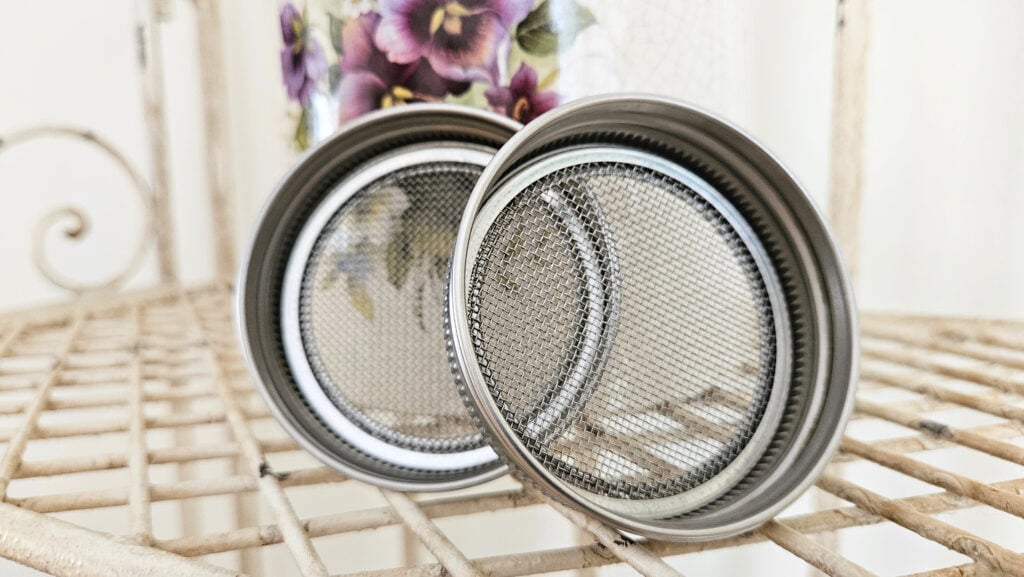Seed sprouting jar lids. Two stainless steel jar lids with a solid rim and fine mesh interior. Sitting on top of a cream colored metal rack. Cream colored pottery with purple painted flowers in the background.