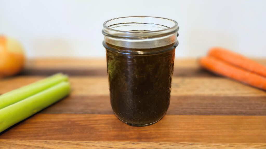 One cup of beef bone broth in a mason jar sitting on top of butcher block. Carrots, celery, and an onion on the butcher block. White wall in the background.