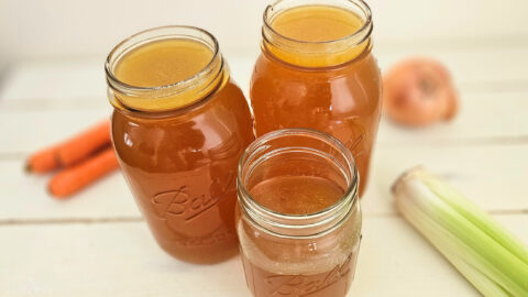Three mason jars filled with amber colored bone broth. Sitting on a cream colored table. Carrots, celery, and an onion on the table.