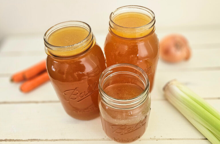 Three mason jars filled with amber colored bone broth. Sitting on a cream colored table. Carrots, celery, and an onion on the table.