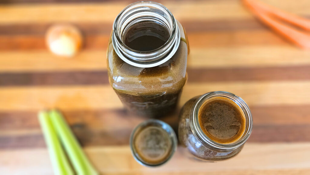 Three jars of beef bone broth sitting on a butcher block counter. Carrots, celery, and an onion on the butcher block.