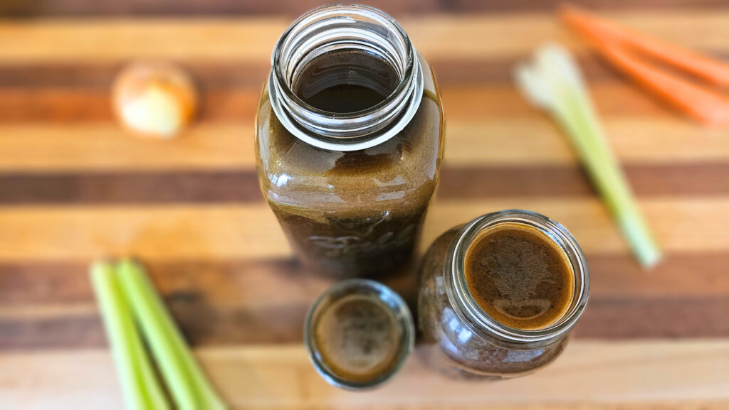 Three different sized mason jars filled with beef bone broth sitting on top of a butcher block countertop. Carrots, celery, and an onion next to the jars.