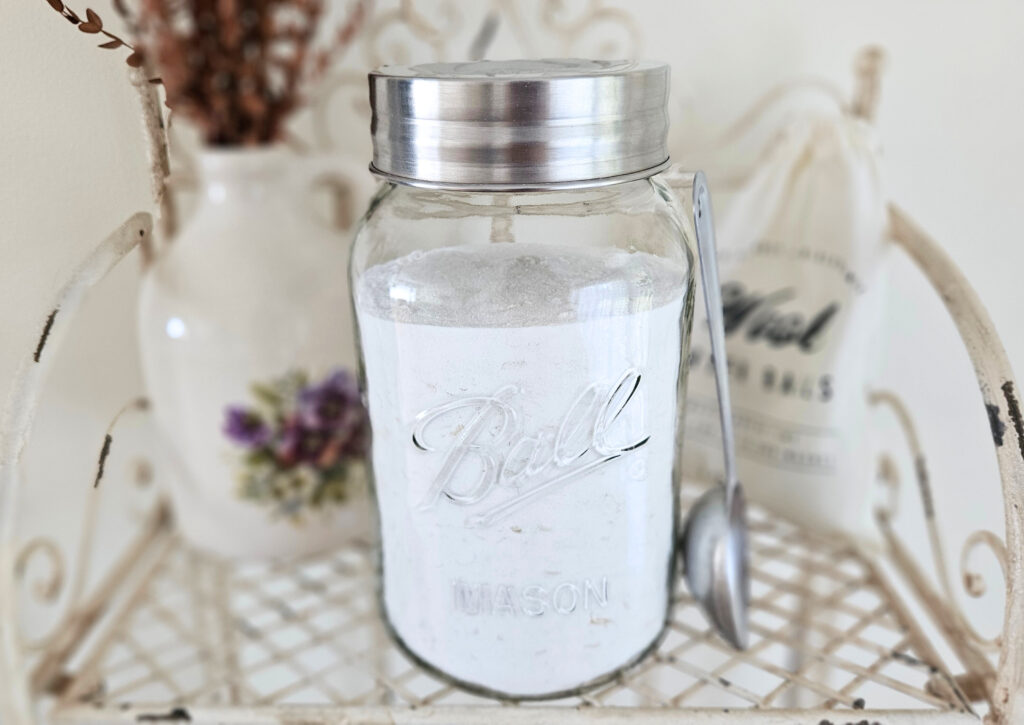 A large glass jar with a metal lid filled with powder laundry detergent. A metal scoop is leaning next to the jar. Pottery filled with dried eucalyptus and a bag of wool dryer balls are in the background.