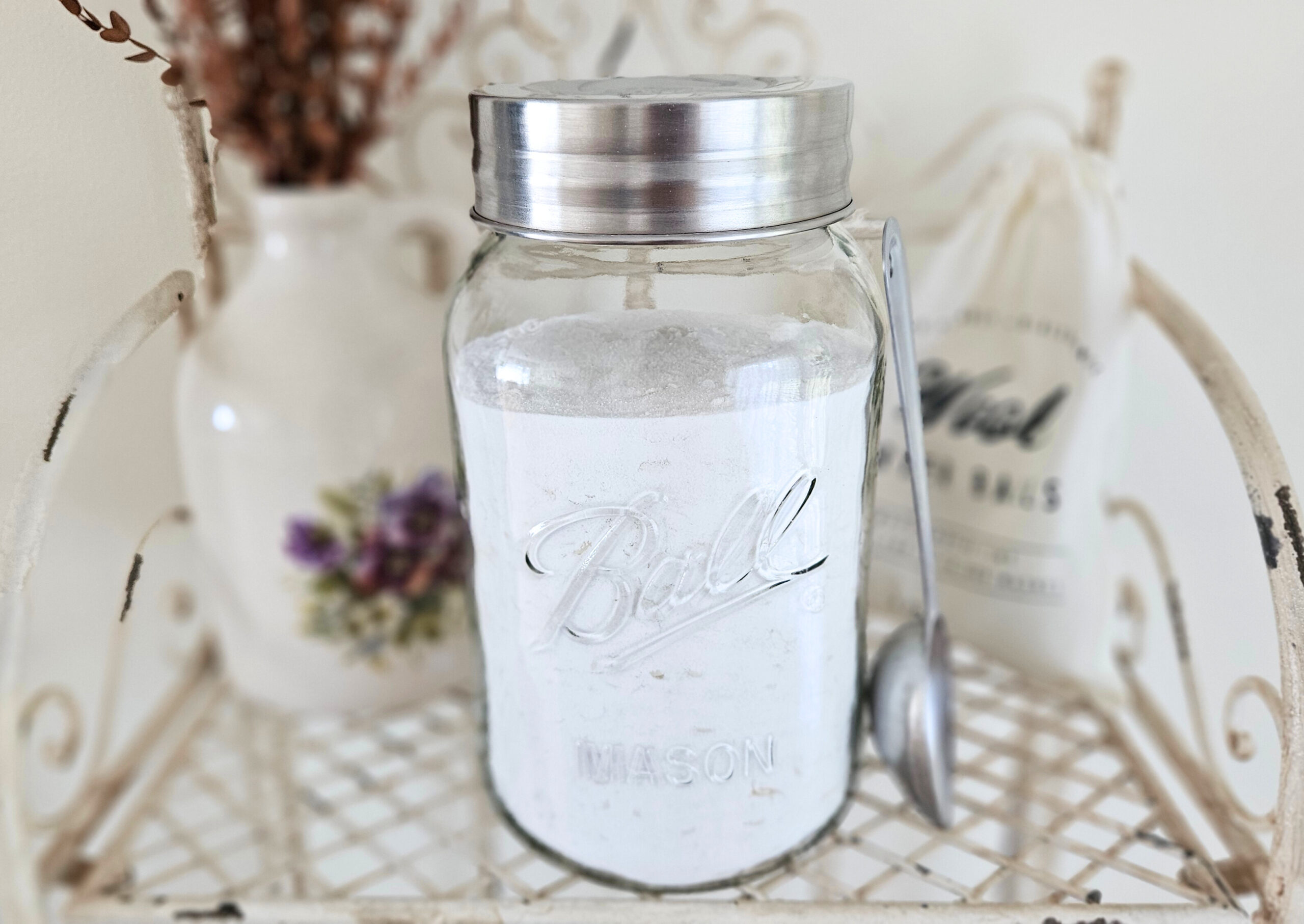 A large glass jar with a metal lid filled with powder laundry detergent. A metal scoop is leaning next to the jar. Pottery filled with dried eucalyptus and a bag of wool dryer balls are in the background.