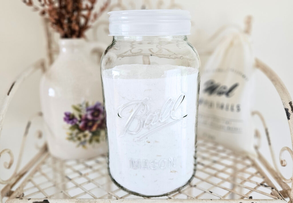 A large glass jar filled with powder laundry detergent. The jar is sitting on top of a metal shelf next to pottery and a bag of wool dryer balls.