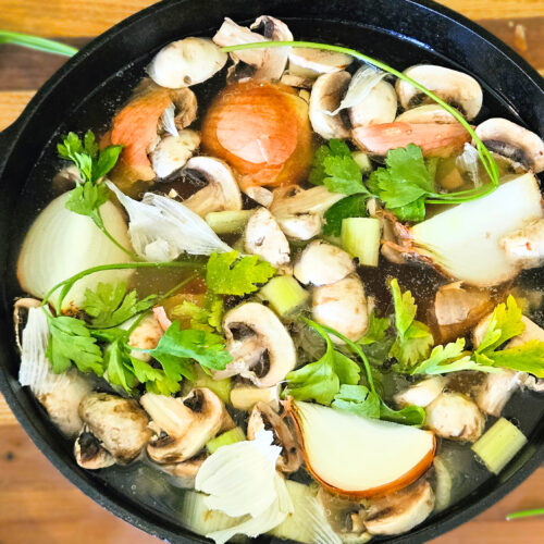 Large cast iron pot filled with beef bones, vegetables, and water, Sitting on butcher block. Carrots, parsley, and garlic on butcher block.