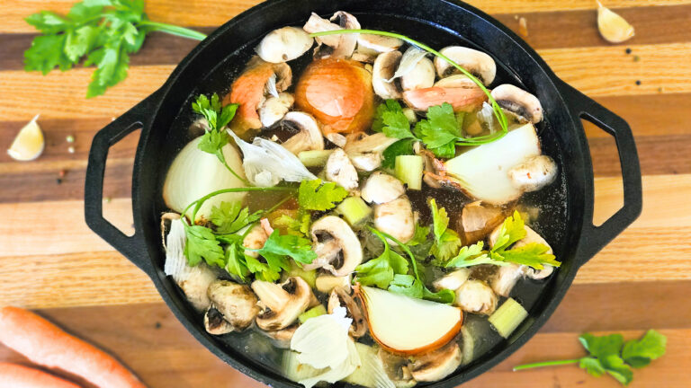 Large cast iron pot filled with beef bones, vegetables, and water, Sitting on butcher block. Carrots, parsley, and garlic on butcher block.